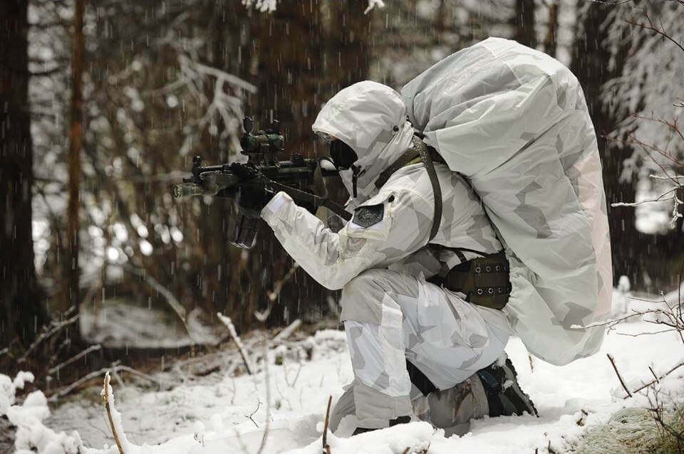Bild av en airsoft spelare ute i skogen, iklädd svenska M90 vinterkamouflage